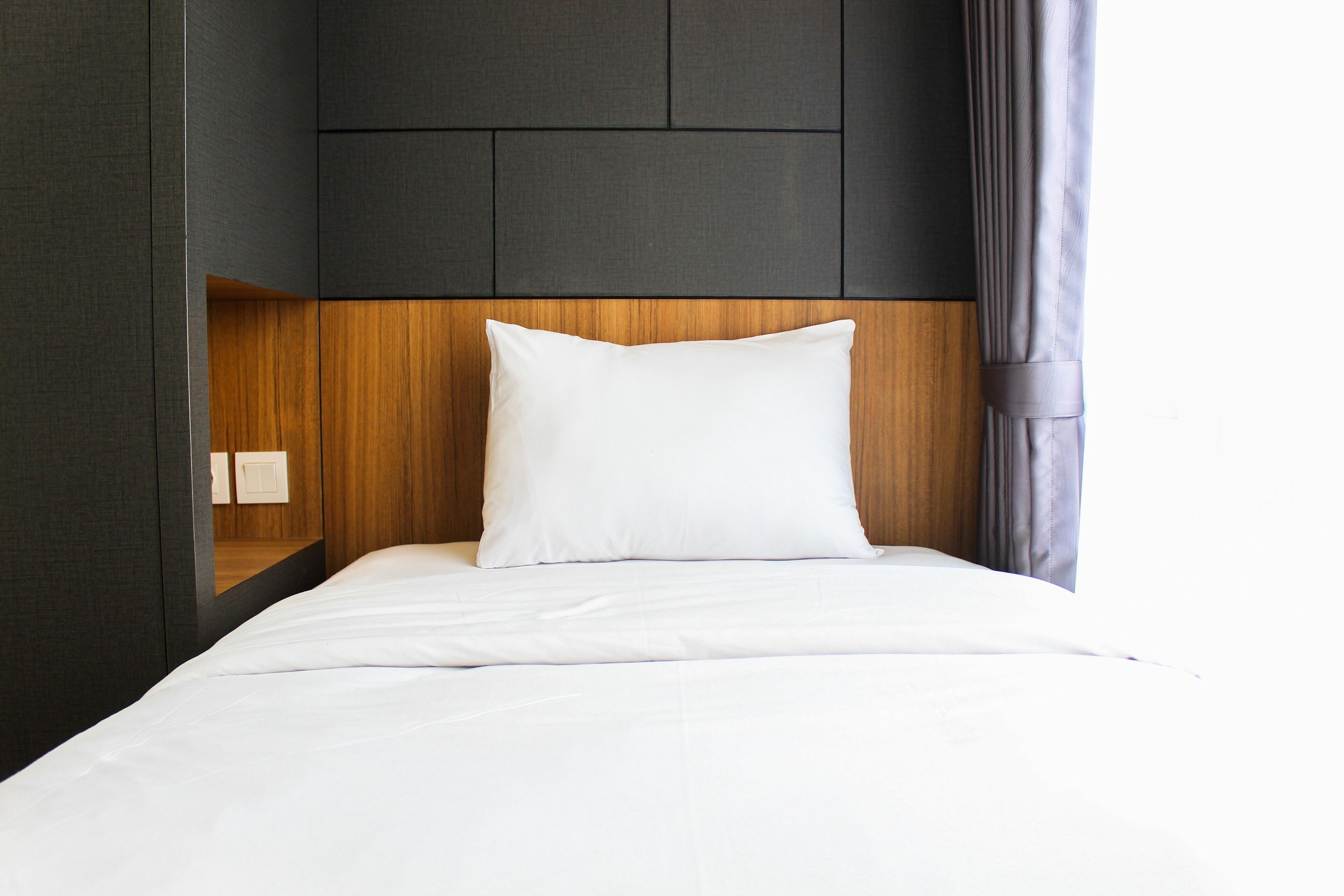 A neatly made bed with a wooden headboard, a white pillow, and a dark wall with a recessed shelf.