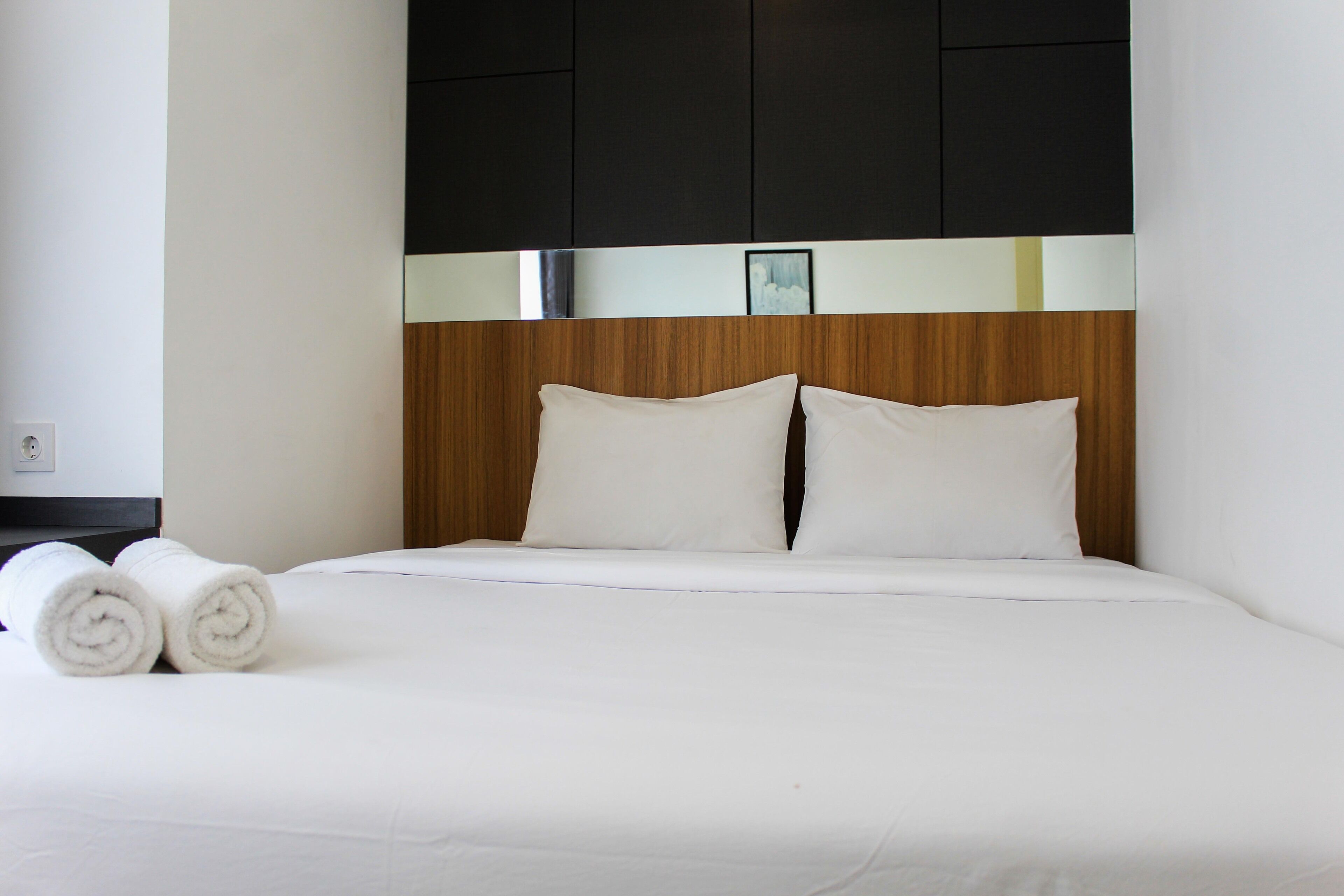 A neatly made bed with white linens and pillows, a wooden headboard with a black and white panel, and two rolled towels on the bed.