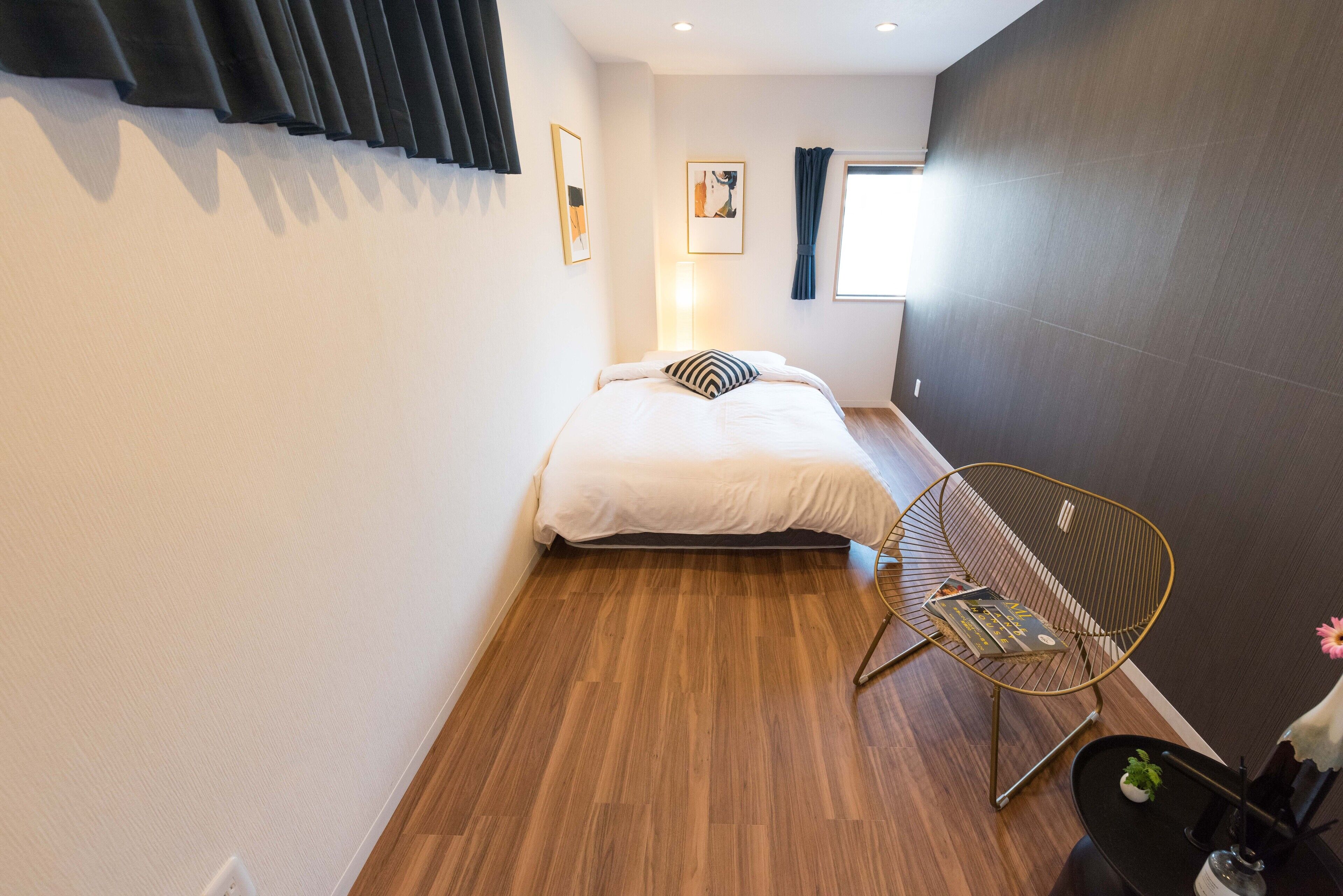 Un dormitorio moderno con piso de madera, una cama con ropa de cama blanca, un almohadón blanco y negro, una silla de alambre dorada y una mesita con una planta.