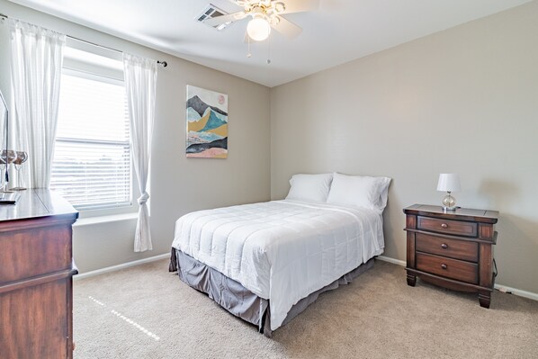 A bedroom with a bed, a nightstand, a dresser, a ceiling fan, and a window with curtains.