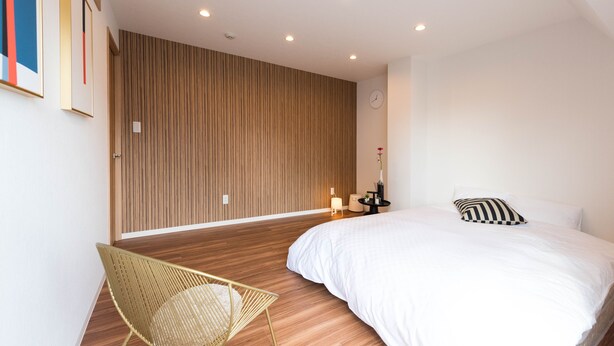 Quarto moderno com piso de madeira, uma cama com roupa de cama branca, uma cadeira, mesa de cabeceira com abajur e um relógio de parede.