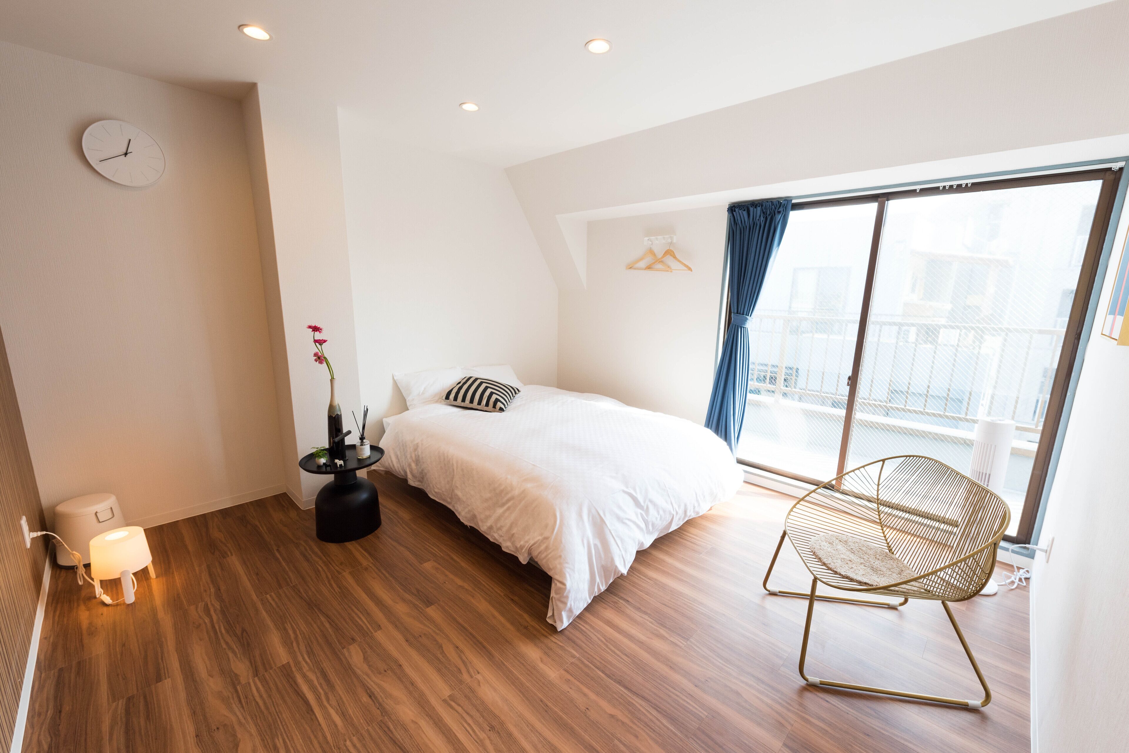 A modern bedroom with a wooden floor, a bed with white bedding, a bedside table with a lamp, and a chair by the window.