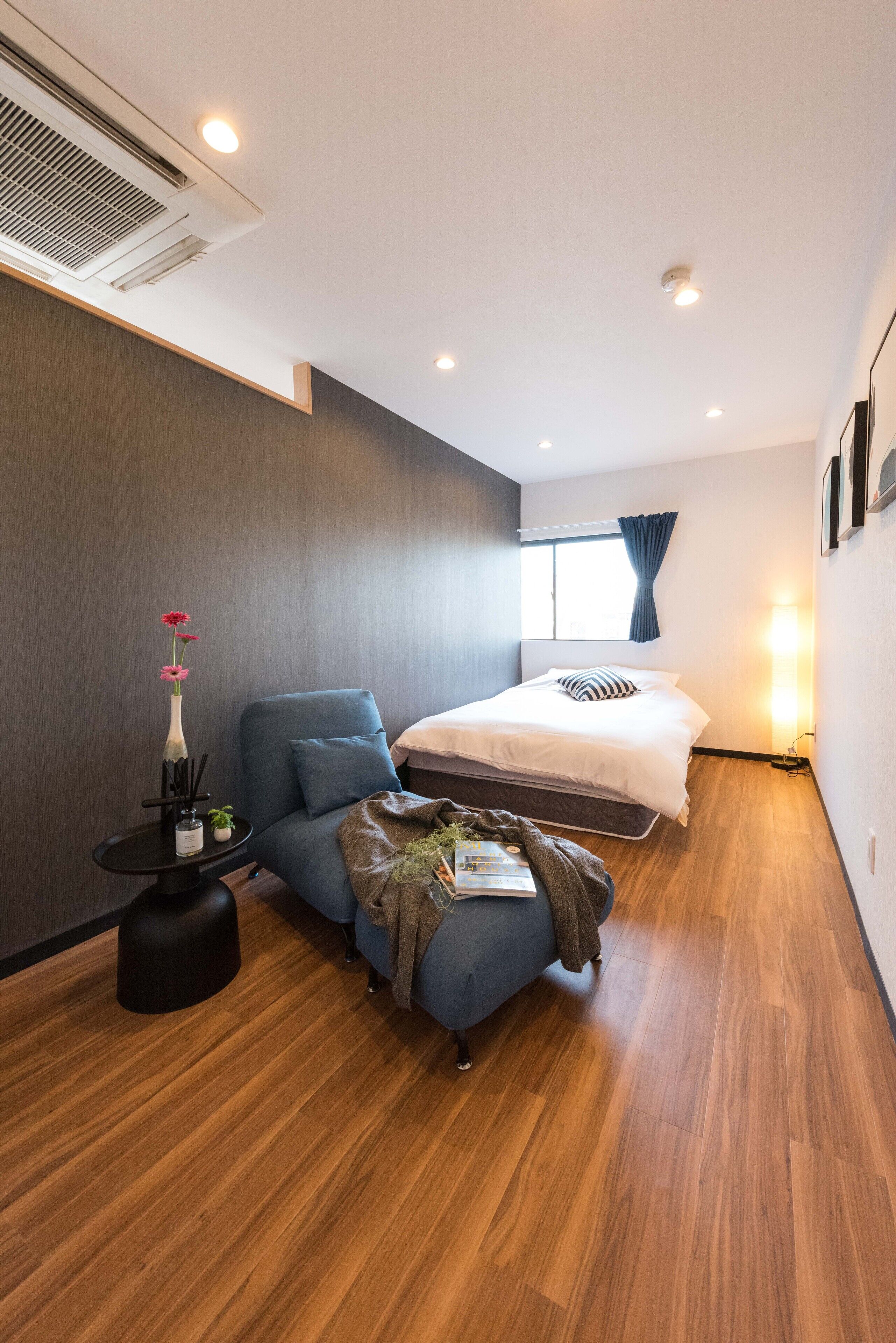 A modern hotel room with a bed, a blue armchair, a small side table with a vase, and a wooden floor.