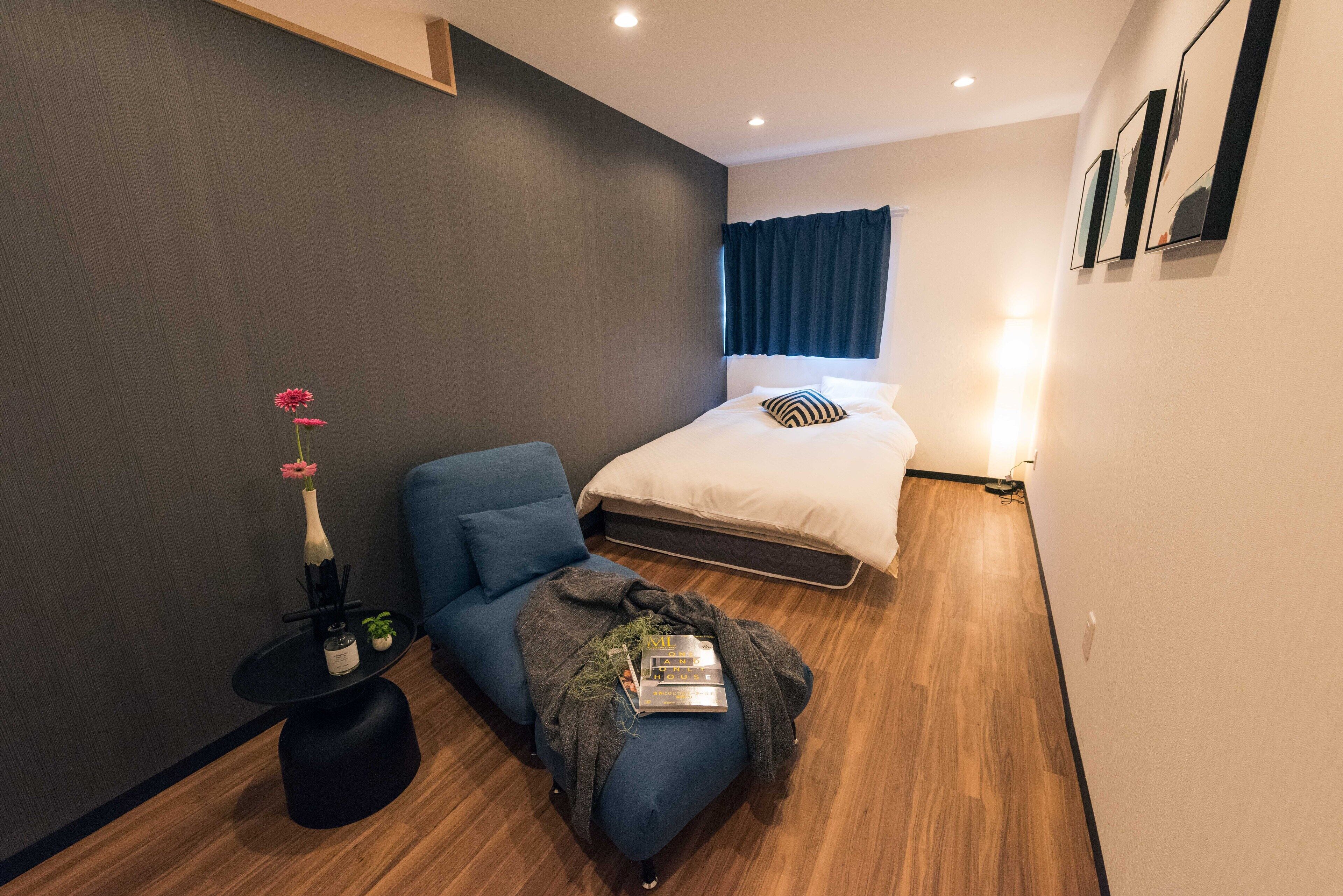 A modern bedroom with a bed, a blue sofa, a small table with a vase and books, and a wooden floor.