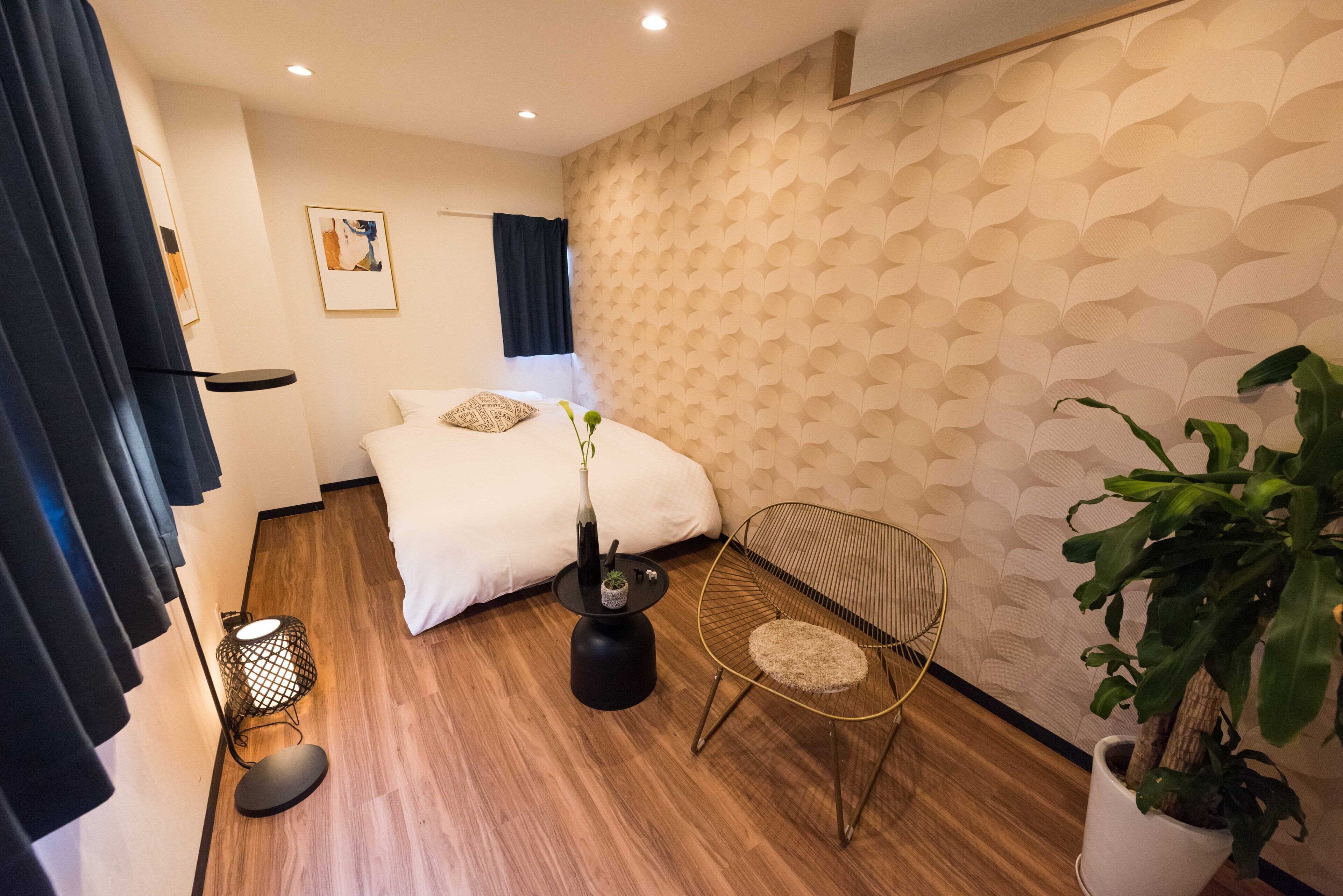 A hotel room with a bed, bedside table, chair, potted plant, and a patterned wall.