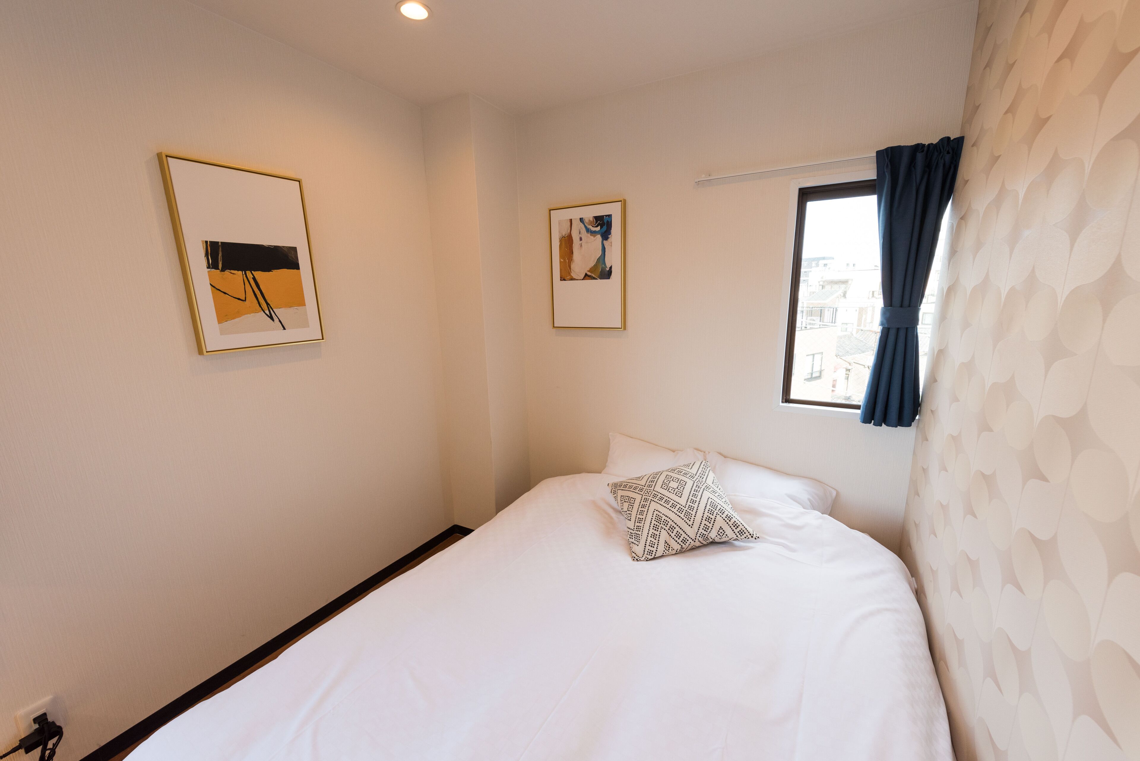 A neatly arranged hotel room with a bed, two framed pictures on the wall, a window with curtains, and a patterned wall.