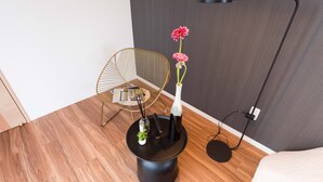 Une pièce moderne avec un plancher en bois, une table ronde noire ornée d’un vase de fleurs, une chaise en fil de fer doré et un lampadaire.