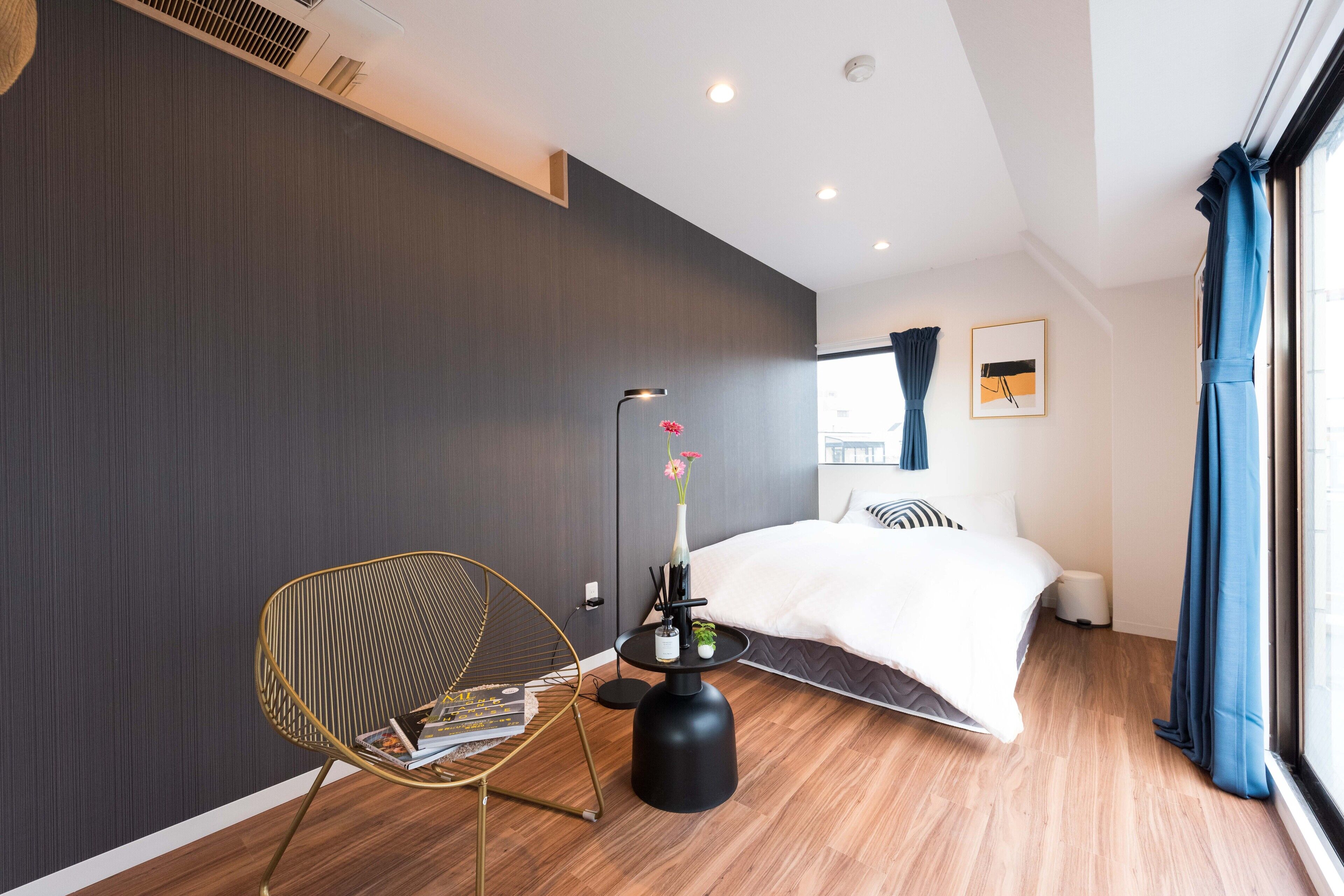A modern hotel room with a large bed, a chair, a small table with a vase, and a window with curtains.