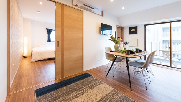 Une chambre d’hôtel moderne avec un plancher en bois, un lit, un coin repas avec une table et des chaises, et une grande fenêtre donnant sur la ville.