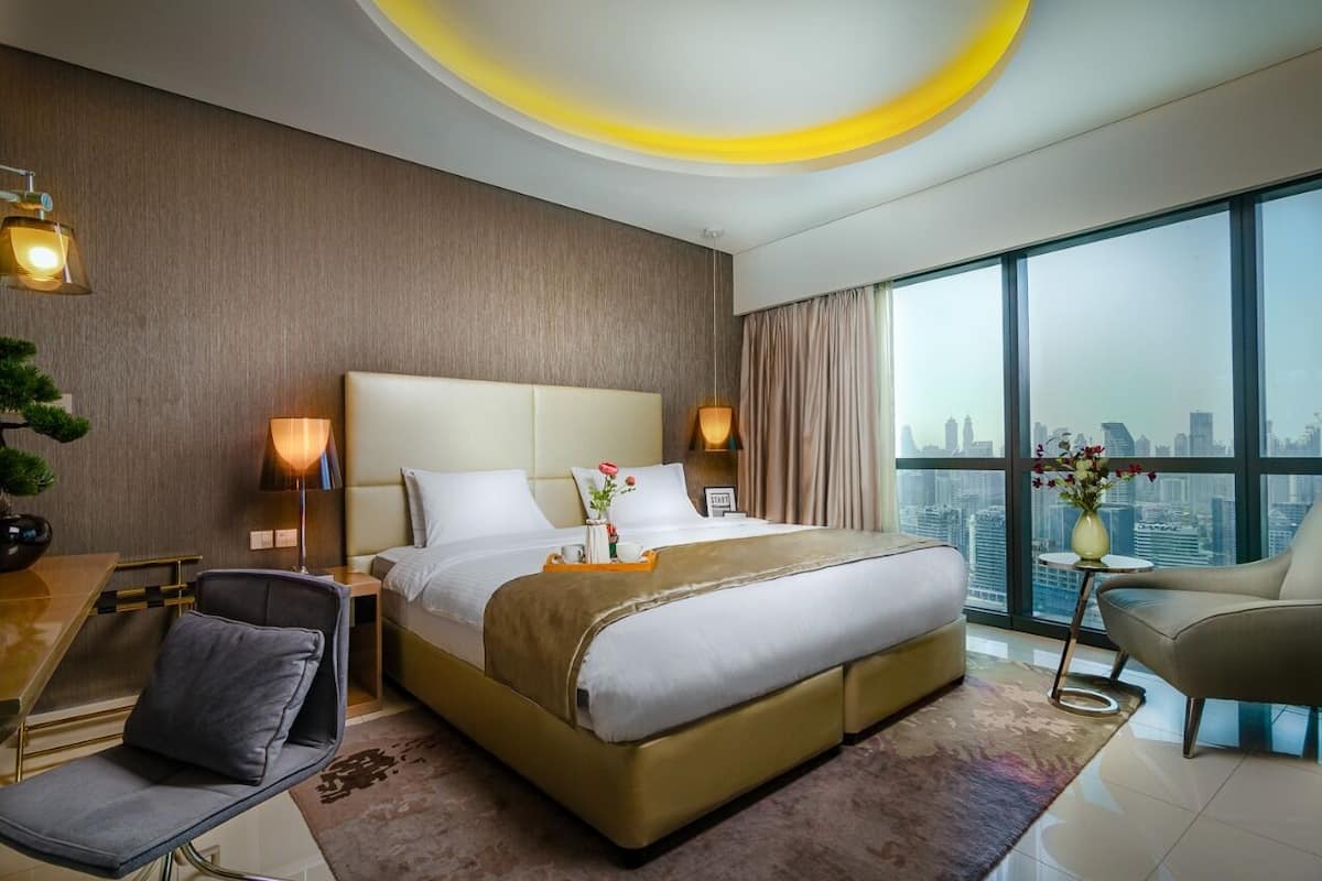 A modern hotel room with a large bed, a desk, a chair, and a view of the city skyline.