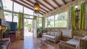 A sunlit living room with stone walls, a television, wicker furniture, and large windows with curtains. - Nice home in Trabia with private swimming pool, can be inside or outside (Trabia)