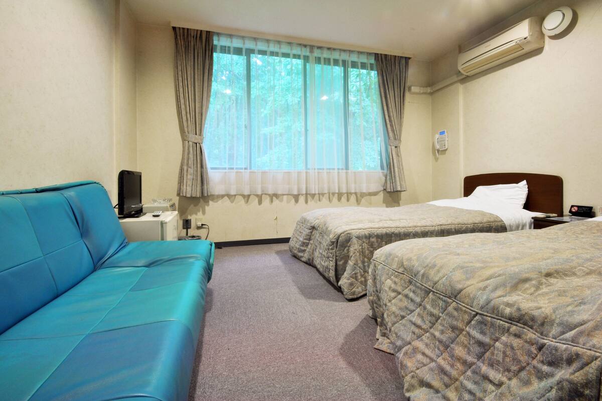 Quarto de hotel com duas camas, um sofá azul, uma janela com cortinas, um ar condicionado e um telefone na parede.