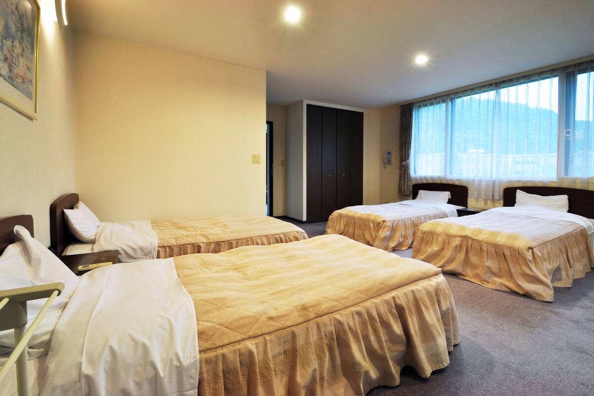 Quarto de hotel com três camas de solteiro, uma mesa de cabeceira com abajur e uma janela grande com cortinas.