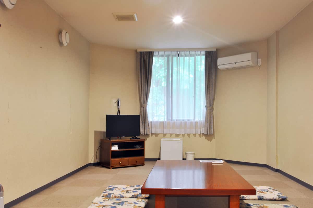 Quarto com uma mesa de madeira, uma televisão sobre um suporte, janela com cortinas e um aparelho de ar condicionado na parede.