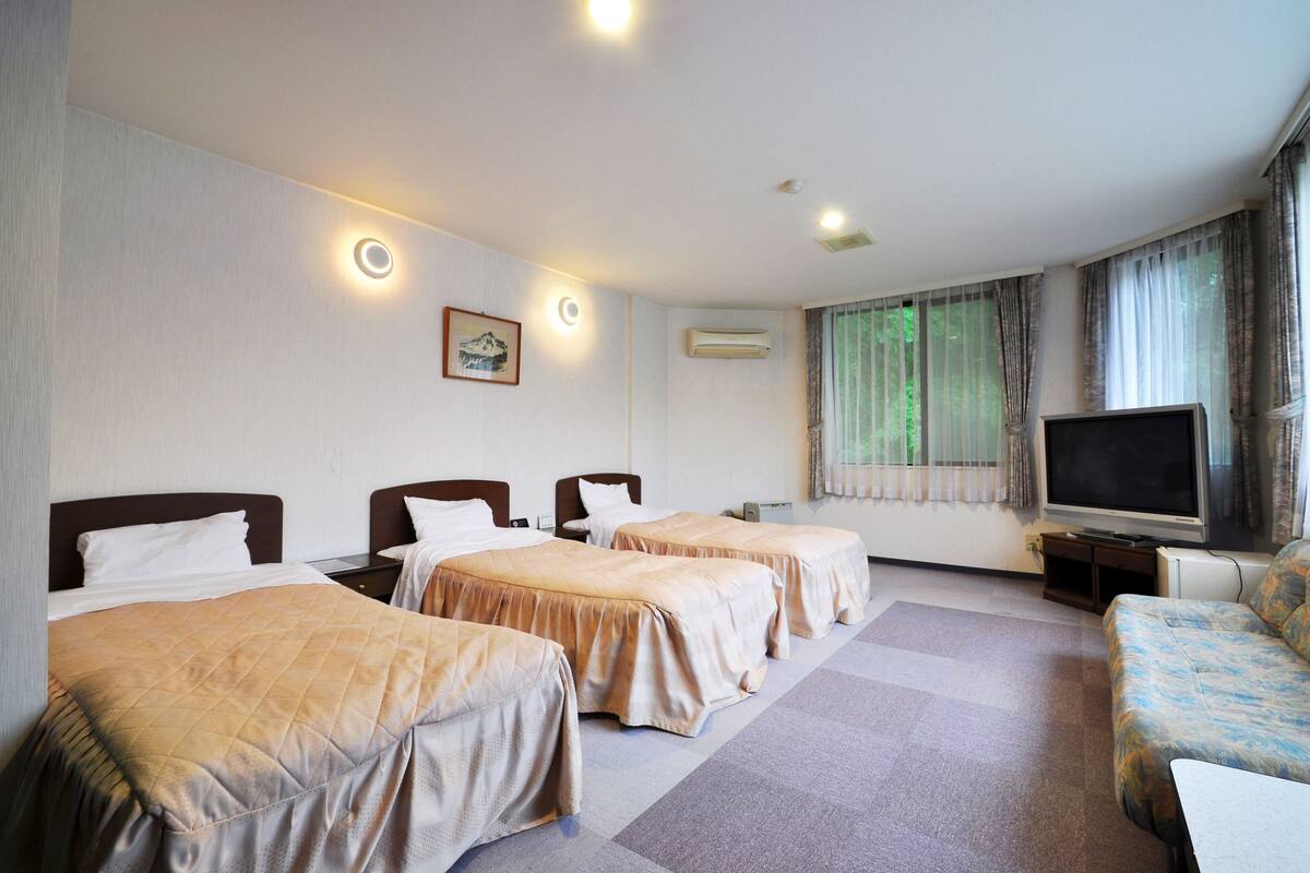 Quarto de hotel com três camas de solteiro, um sofá, uma televisão e uma janela com cortinas.