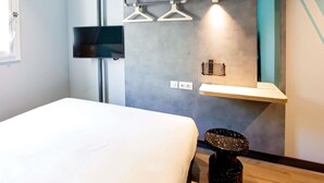 Una habitación de hotel moderna con cama, televisor montado en la pared, estante con perchas y una mesita con un objeto decorativo.