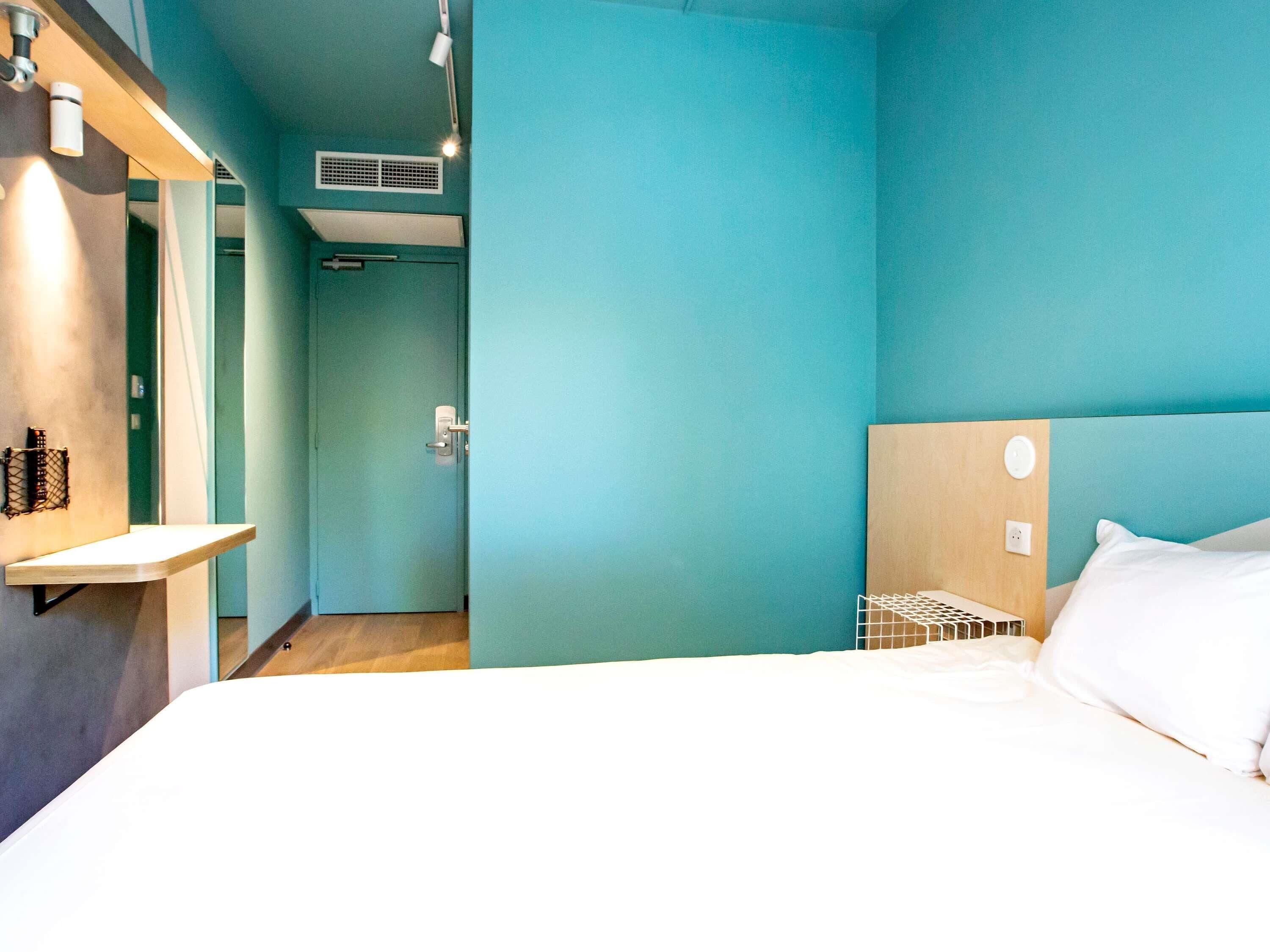 Una habitación de hotel moderna con paredes verde azulado, un cabecero de madera, una cama blanca y una mesita de noche pequeña.
