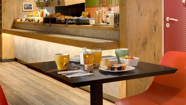 Un área de desayuno con mostrador de madera que exhibe pasteles y bebidas, una mesa con alimentos para el desayuno y una pared con estampado de lunares rojos.