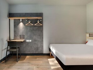 A modern hotel room with a bed, a desk with a chair, and a wall-mounted coat rack.