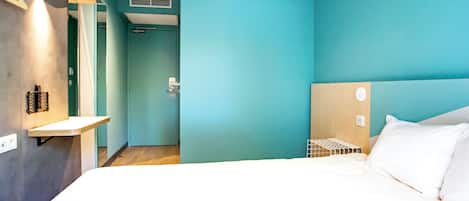 A modern hotel room with teal walls, a wooden headboard, a white bed, and a small shelf with toiletries.
