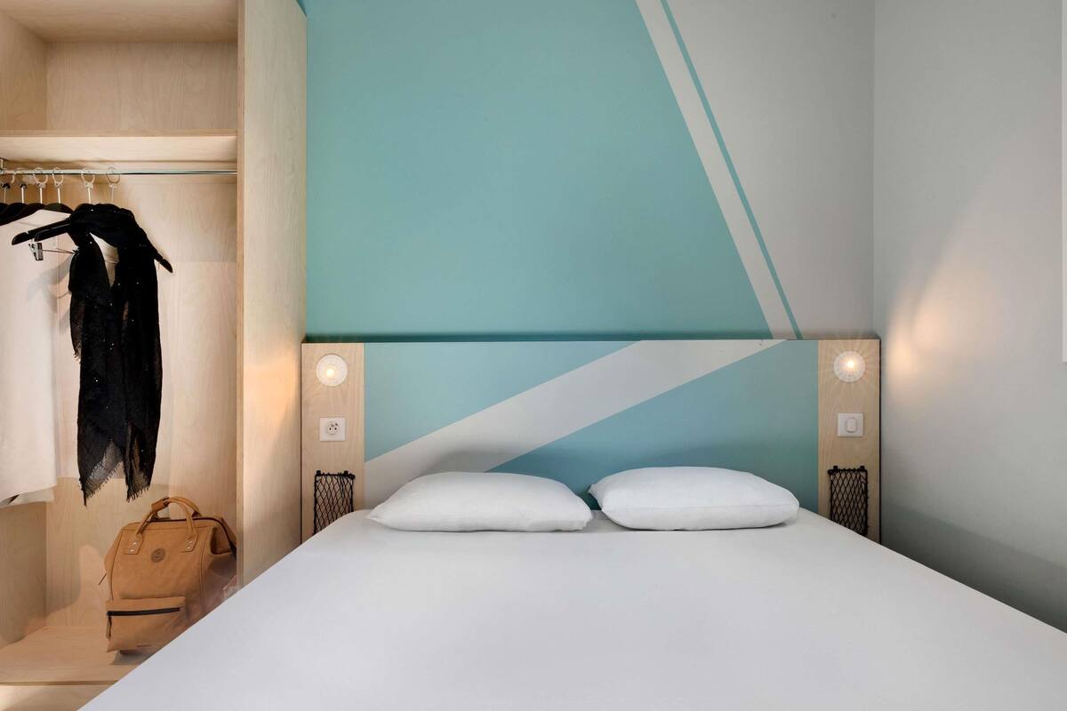Une chambre d’hôtel moderne comprenant un lit, deux tables de chevet, un porte-manteau et un banc en bois.
