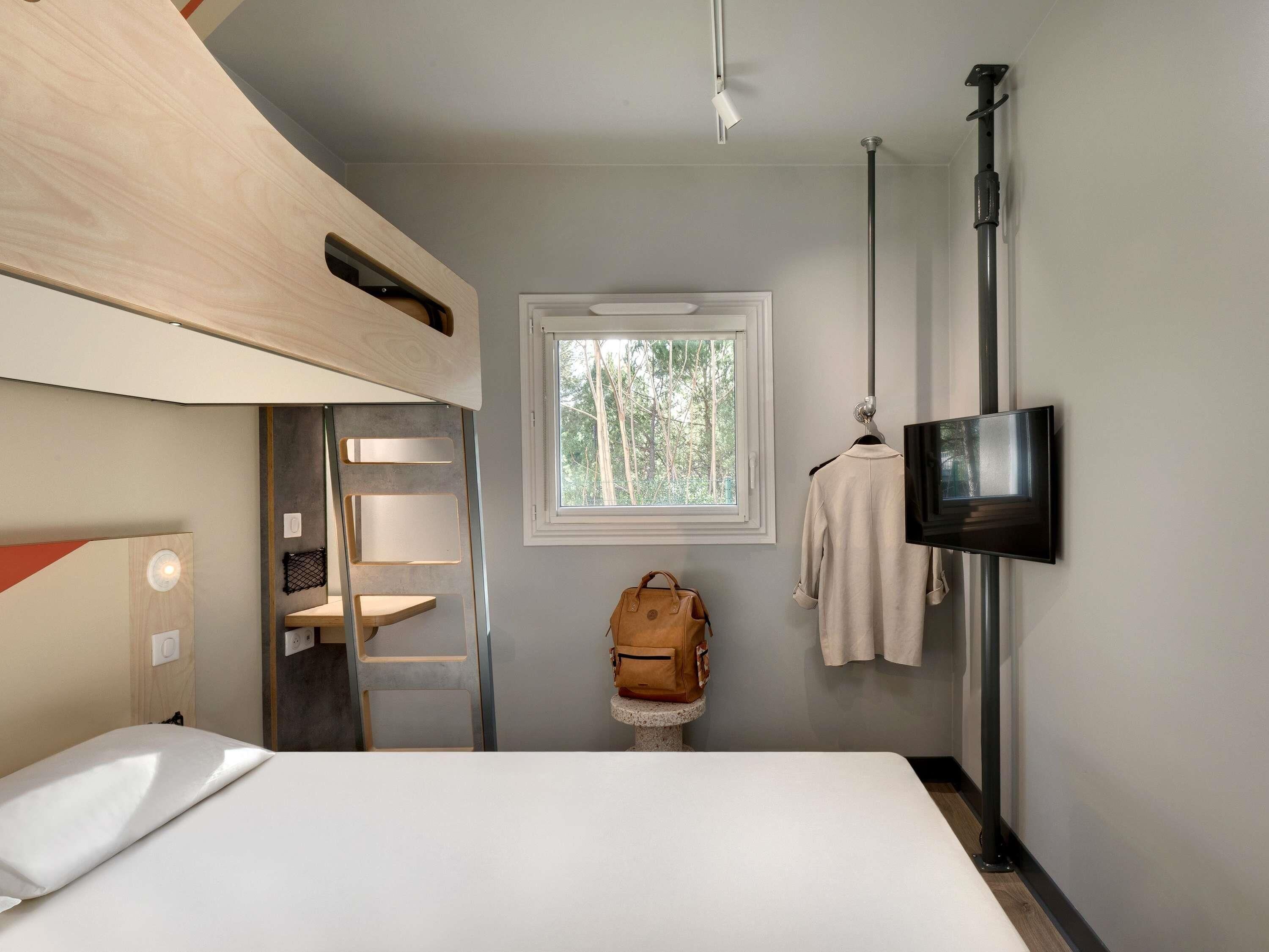A compact room with a bunk bed, a TV mounted on the wall, a window with a view of trees, and a ladder.