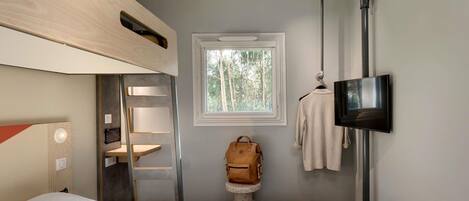 A compact room with a bunk bed, a TV mounted on the wall, a window with a view of trees, and a ladder.