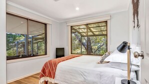 Un dormitorio con una cama grande, un escritorio y una silla. Hay dos ventanas, una con vista a árboles y la otra a una edificación.