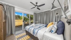 A bedroom with a bed, a ceiling fan, a desk lamp, and a view through a sliding glass door to an outdoor area. - Akora House at Hyams Beach (Hyams Beach)