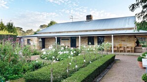 A stone building with a covered porch, surrounded by a well-maintained garden with flowering plants and a gravel path. - Aylmerton House + Cottage for 14, Southern Highlands (Mittagong)
