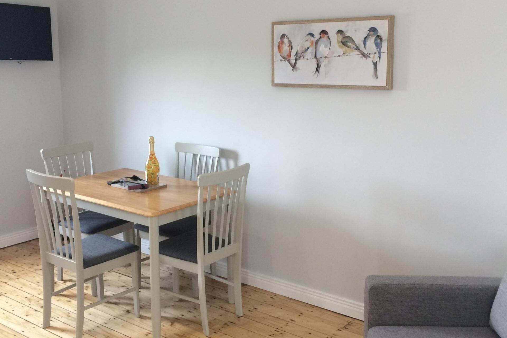 A dining area with a wooden table and white chairs, a painting of birds on the wall, and a grey sofa.