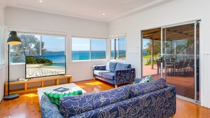 A spacious living room with a large sofa, a coffee table, and a television. There are large windows offering a view of the beach and the sea. - Bimbadgen at Hyams Beach (HYAMS BEACH)