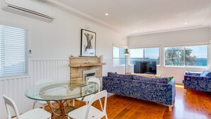 A modern living room with a glass dining table, white chairs, a blue patterned sofa, and a fireplace. - Bimbadgen at Hyams Beach (HYAMS BEACH)