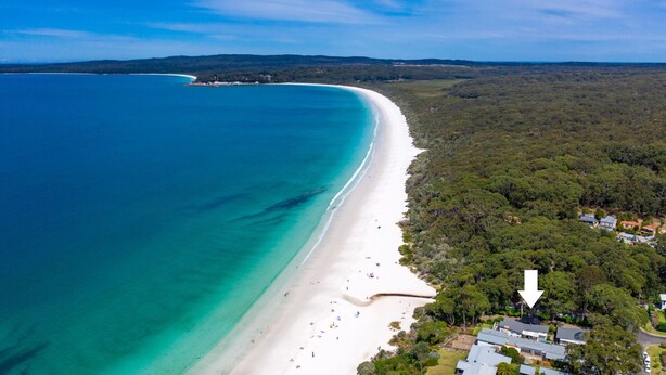An aerial view of a coastal area with a long sandy beach, dense forest, and a few buildings near the shore.