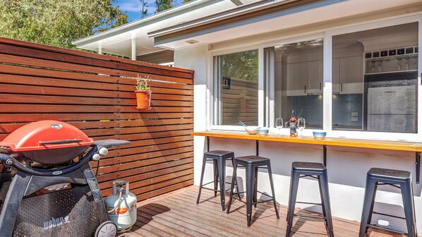 An outdoor patio with a barbecue grill, stools, and a table with glasses and a bottle. - Black Wattle at Hyams Beach (Hyams Beach)