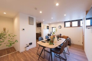 Ein moderner Essbereich mit einem Holztisch und Stühlen, im Hintergrund eine Küche und ein Blick ins Freie durch ein Fenster.