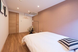 Ein Schlafzimmer mit Holzboden, einem Bett mit weißer Bettwäsche, einem schwarz-weiß gestreiften Kissen, einer Stehlampe und einer Wand-Klimaanlage.