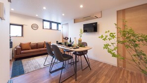 A modern living room with a brown leather sofa, a dining table with chairs, a flat-screen TV mounted on the wall, and a wooden floor. - TORA HOTEL Skytree B (Tokyo)