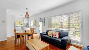 A modern living room with a black leather sofa, wooden dining table, and chairs. There is a kitchen area with white cabinets and a window with blinds.
