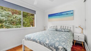 A bedroom with a bed, a nightstand, a lamp, a window with a view of trees, and a painting of a beach.