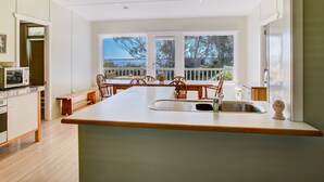 A modern kitchen with a large island, wooden chairs, and a view of the outdoors through large windows. - Calypso's Cottage at Hyams Beach (Hyams Beach)
