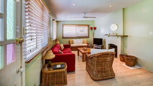 A cozy living room with a red sofa, wicker furniture, a television, and a fireplace. - Calypso's Cottage at Hyams Beach (Hyams Beach)