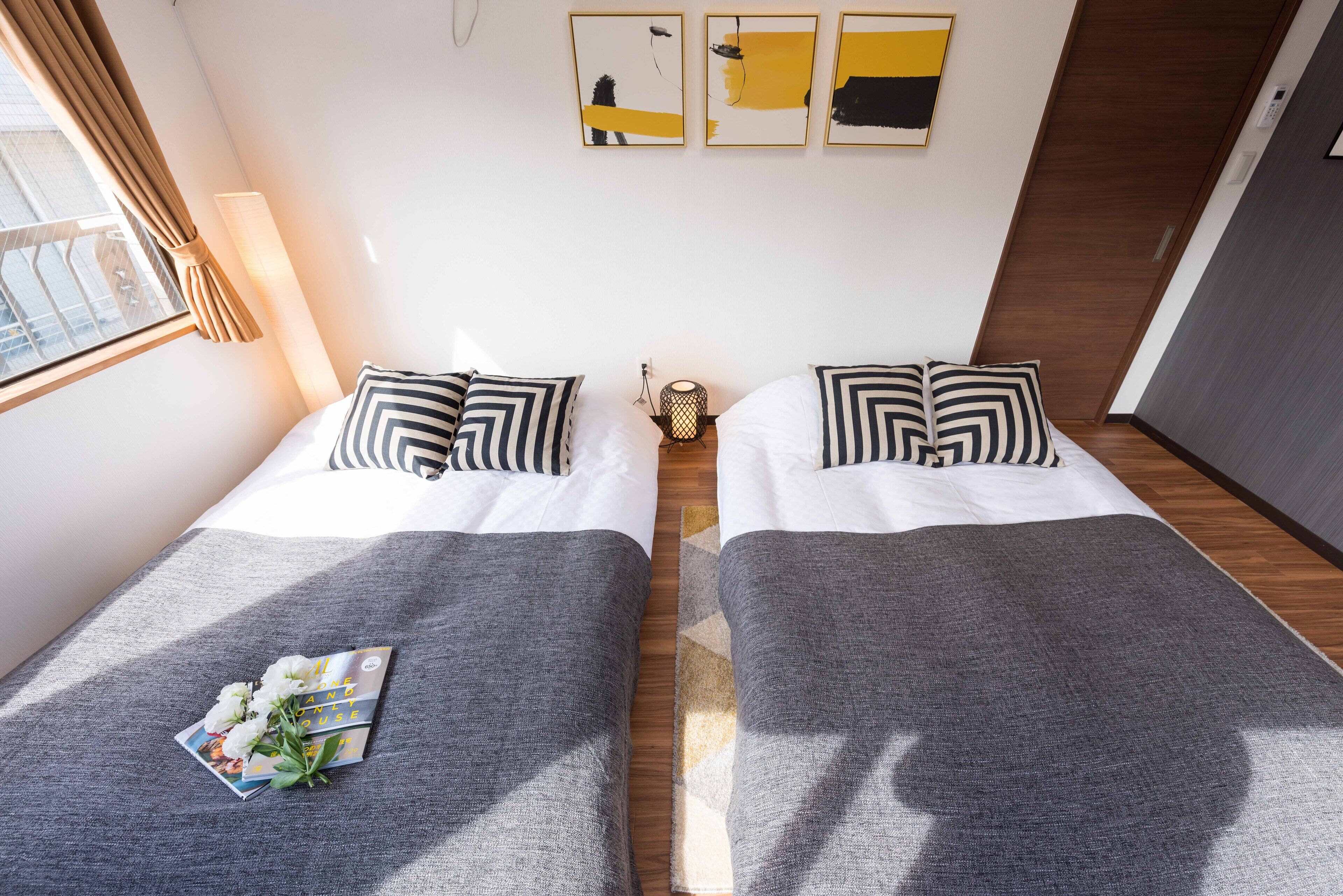 A hotel room with two beds, a window with curtains, a lamp, and artwork on the wall.