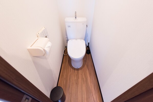 A small, narrow bathroom with a white toilet, a paper towel dispenser, and a wooden floor.
