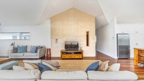 Amplia sala de estar con techo alto, un televisor grande sobre un mueble de madera, un conjunto de sofás y una zona de cocina con refrigerador.