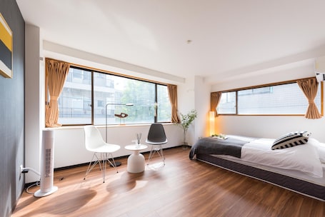A modern hotel room with a bed, a desk, two chairs, a standing heater, and large windows with curtains.