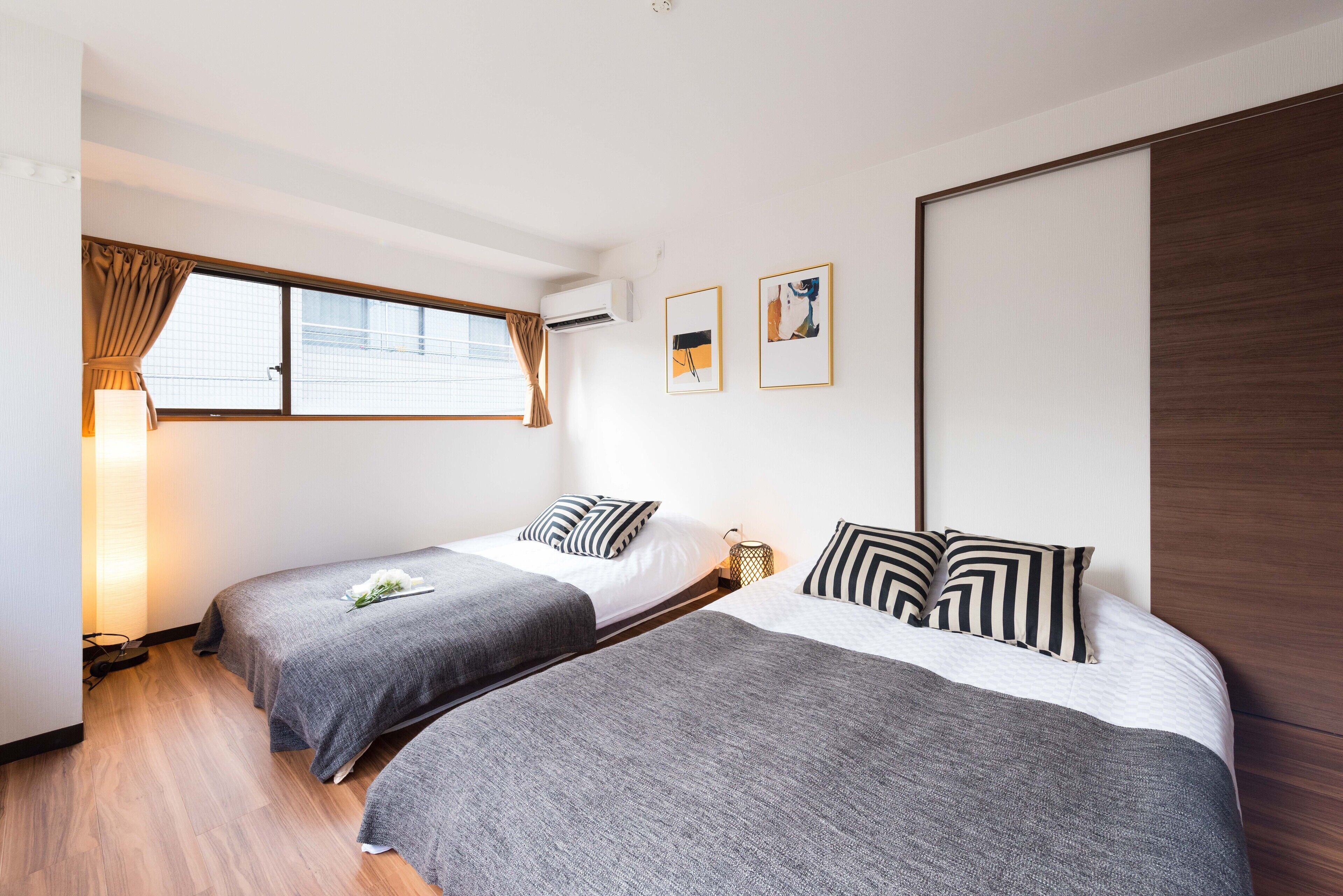 A hotel room with two single beds, a window with curtains, an air conditioning unit, and two framed pictures on the wall.