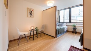 A modern room with a wooden floor, a bed with a blue headboard, a small table with a plant, and a white chair. - TORA Hotel Ueno 193D (Tokyo)