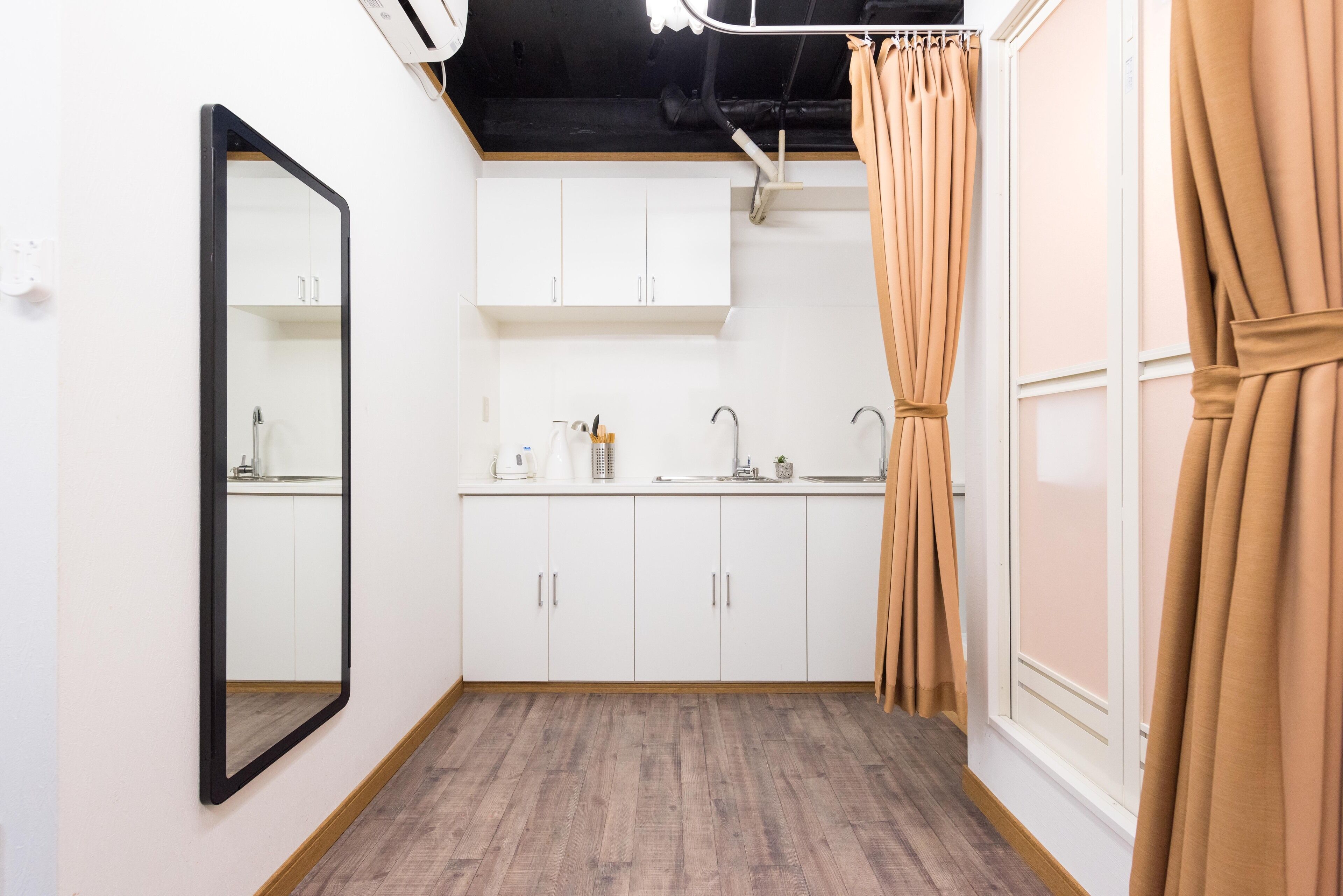 Una habitación pequeña y limpia con armarios blancos, un espejo, un lavamanos y una ventana con cortinas color beige.