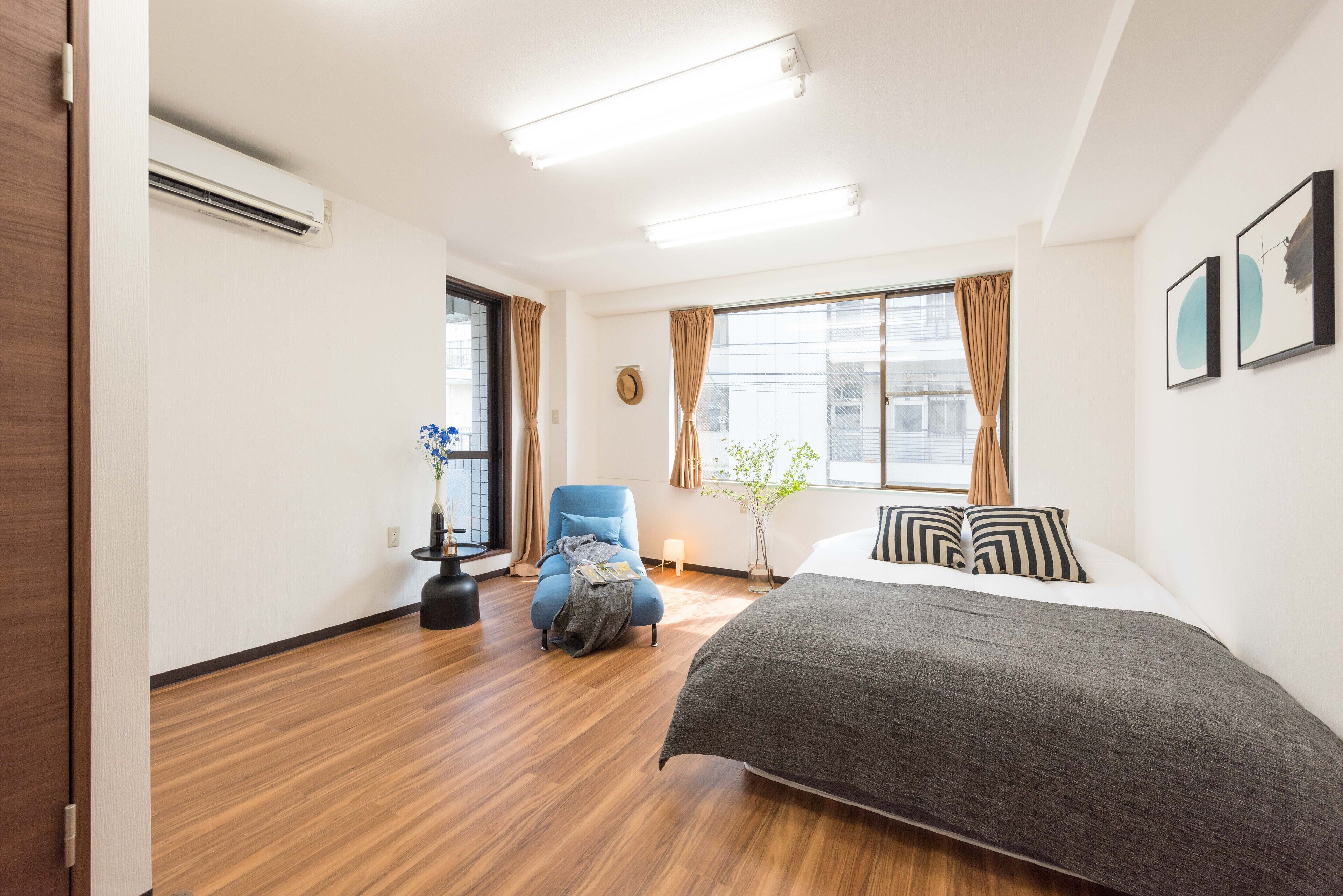 Une chambre d’hôtel moderne avec un plancher en bois, un lit recouvert d’une couverture grise, un fauteuil bleu, une petite table avec un vase et une fenêtre avec des rideaux.